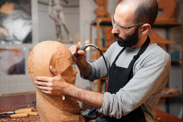 Crop male woodworker in apron and protective glasses carving wooden sculpture with professional engraver tool while working in light joinery - ADSF32461