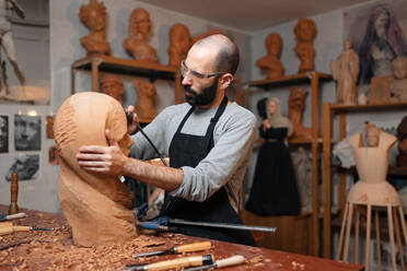 Crop male woodworker in apron and protective glasses carving wooden sculpture with professional engraver tool while working in light joinery - ADSF32462