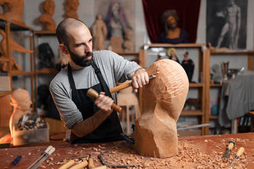 Serious bearded carpenter shaping round wooden detail with professional mallet and chisel while working in joinery with special equipment - ADSF32464