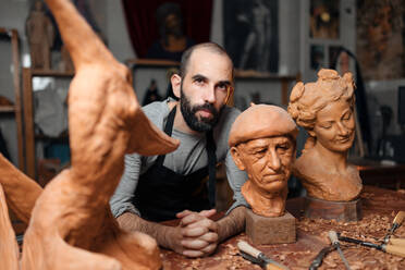 Pensive bearded male woodworker in apron standing at table with carved wooden sculptures and professional instruments during work in joinery - ADSF32465