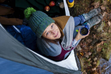 Smiling woman wrapped in blanket holding coffee mug inside tent - MRRF01735