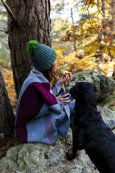 Frau mit Strickmütze isst Apfel mit Hund auf Felsen im Herbstwald - MRRF01736