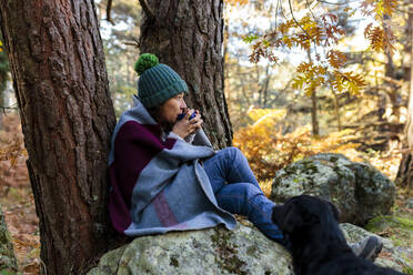 Frau in Decke eingewickelt, Kaffee trinkend, auf einem Felsen sitzend, neben einem Hund im Wald - MRRF01737