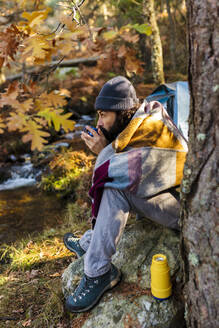 Mann trinkt Kaffee auf einem Felsen im Herbstwald sitzend - MRRF01742