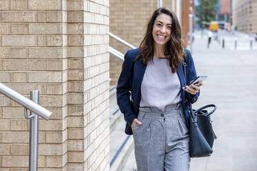 Smiling businesswoman with hand in pocket holding mobile phone walking on footpath - WPEF05580