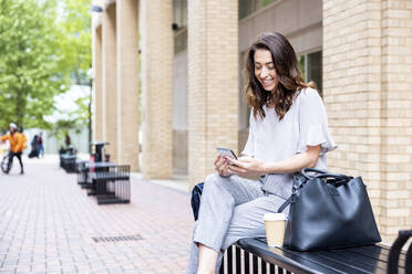 Smiling businesswoman using smart phone sitting on bench with bag and disposable cup - WPEF05588