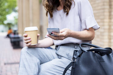 Geschäftsfrau mit Smartphone auf einer Bank sitzend, mit Einwegbecher und Tragetasche - WPEF05589