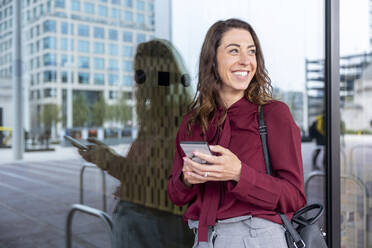 Happy businesswoman holding mobile phone leaning on glass wall in city - WPEF05599