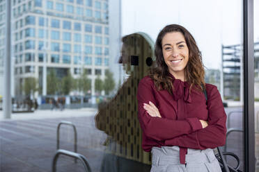 Smiling businesswoman with arms crossed leaning on glass wall in city - WPEF05600