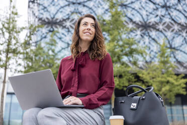 Nachdenkliche Geschäftsfrau mit Laptop und Tasche, die im Büropark sitzt - WPEF05603