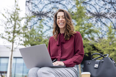 Geschäftsfrau sitzt mit Laptop lachend im Büropark - WPEF05604