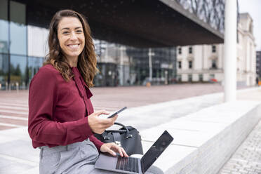 Lächelnde Geschäftsfrau mit Laptop und Smartphone sitzt auf einer Bank im Büropark - WPEF05606