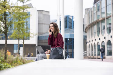 Smiling businesswoman with laptop talking through mobile phone sitting on bench at office park - WPEF05608