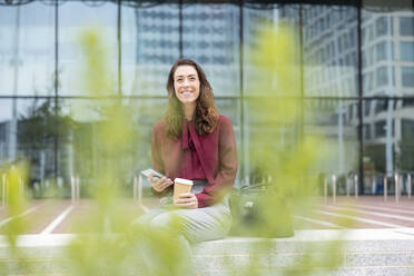 Smiling businesswoman holding mobile phone and disposable cup sitting at office park - WPEF05610