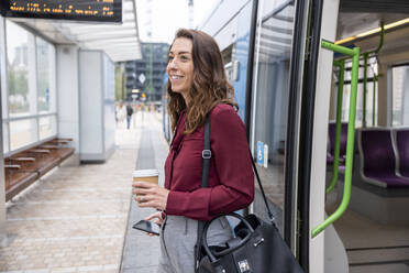 Lächelnde Geschäftsfrau mit Einwegbecher und Mobiltelefon beim Aussteigen aus dem Zug am Bahnhof - WPEF05612