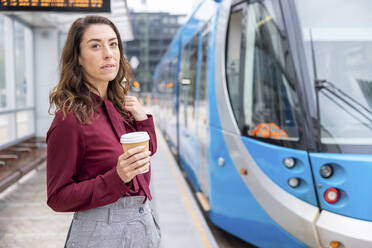 Businesswoman holding disposable cup waiting for train at railroad station - WPEF05613