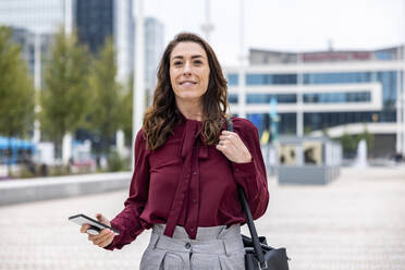 Lächelnde Geschäftsfrau mit Smartphone auf dem Fußweg in der Stadt - WPEF05618