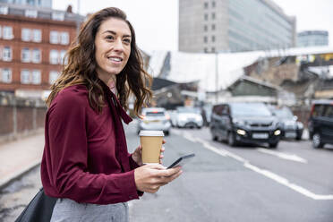 Lächelnde Geschäftsfrau, die auf ein Taxi wartet, mit Handy und Kaffeetasse auf einer Stadtstraße - WPEF05621