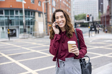 Lächelnde Geschäftsfrau, die über ihr Smartphone spricht, auf einer Straße in der Stadt - WPEF05623