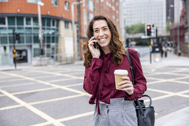 Smiling businesswoman talking through smart phone standing on city street - WPEF05623