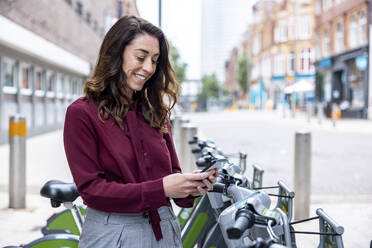 Lächelnde Geschäftsfrau, die ein Mobiltelefon benutzt, um ein Fahrrad für den Arbeitsweg in der Stadt zu mieten - WPEF05625