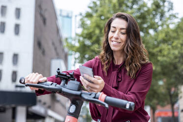 Smiling businesswoman using mobile phone to unlock electric bicycle in city - WPEF05626