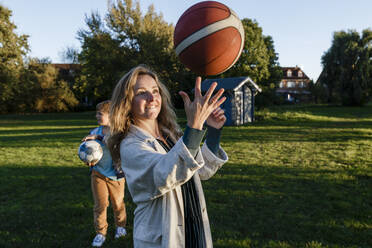 Frau dreht Ball auf Finger mit Sohn im Hintergrund im Park - IHF00703