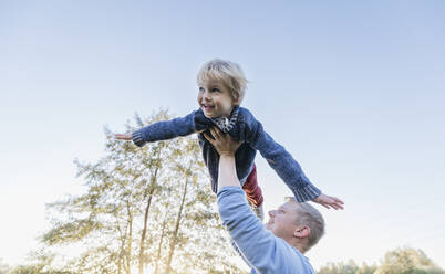 Smiling man holding aloft blond boy flying under clear sky - IHF00707