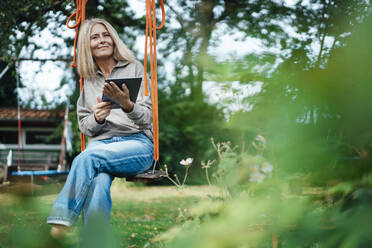 Blond woman with tablet PC sitting on swing at backyard - JOSEF06103