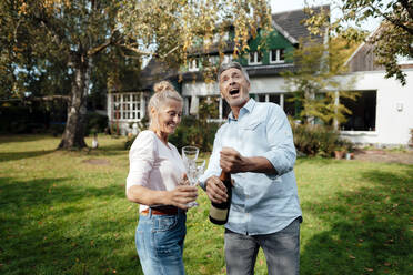 Cheerful man opening champagne by woman at backyard - JOSEF06123