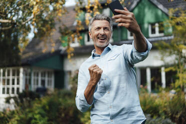 Happy man cheering and taking selfie through smart phone at backyard - JOSEF06151