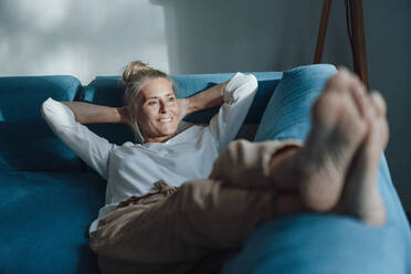Lächelnde Frau mit Händen hinter dem Kopf entspannt auf dem Sofa - JOSEF06163