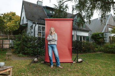 Woman with arms crossed in front of red backdrop at backyard - JOSEF06179