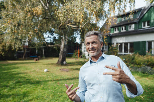 Smiling man gesturing hand sign at backyard - JOSEF06190