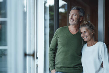 Smiling couple at home looking out of window - JOSEF06232