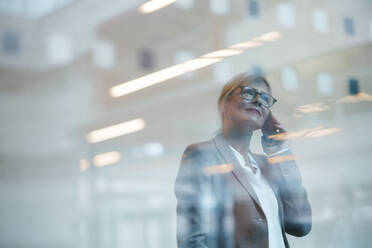 Geschäftsfrau, die mit einem Mobiltelefon spricht, gesehen durch ein Glasfenster im Büro - JOSEF06356