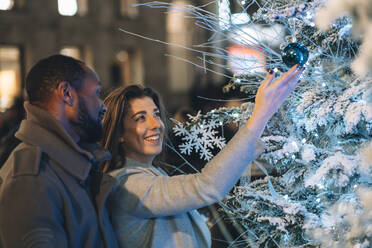 Lächelnde Frau berührt Weihnachtsschmuck am Baum durch Mann in der Nacht - ASGF01857