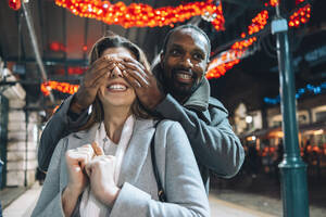 Mann verdeckt Augen seiner Freundin auf beleuchtetem Weihnachtsmarkt - ASGF01859