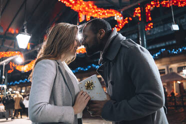 Man with gift box rubbing woman's nose at Christmas market - ASGF01861