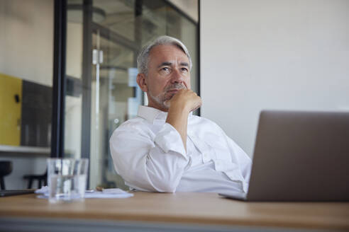 Älterer Geschäftsmann mit Laptop auf dem Tisch, der im Büro nachdenkt - RBF08382