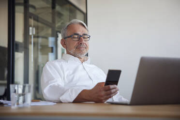 Lächelnder Geschäftsmann, der ein Mobiltelefon im Büro benutzt - RBF08385