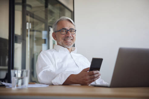Lächelnder reifer Geschäftsmann mit Smartphone und Laptop im Büro - RBF08386