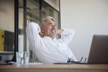 Happy businessman sitting with hands behind head at work place - RBF08397