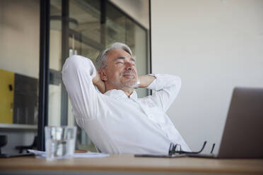 Smiling businessman relaxing with hands behind head at office - RBF08398