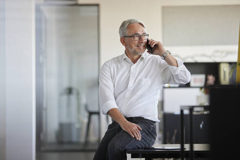 Lächelnder Geschäftsmann, der im Büro mit seinem Handy telefoniert - RBF08406