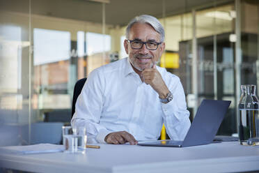 Smiling businessman with laptop sitting at table in office - RBF08420