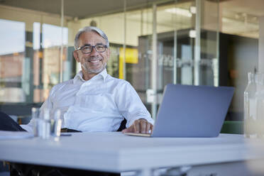 Happy mature businessman with laptop at table in office - RBF08421