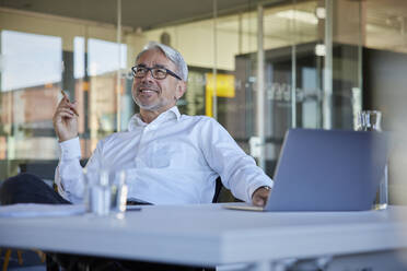 Smiling businessman contemplating while using laptop in office - RBF08423