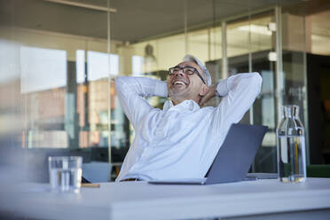 Happy businessman sitting with hands behind head in office - RBF08425