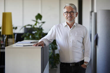 Smiling businessman standing with hand in pocket at office - RBF08433